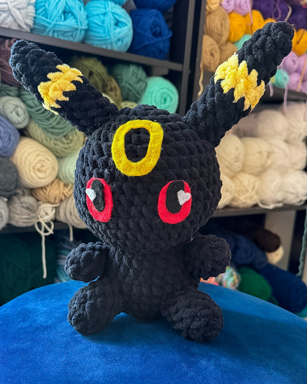 A black crocheted plush toy resembling a small fantasy creature with yellow ear patches, red circular eyes with white heart centers, and a yellow ring on the forehead, sitting on a blue surface with shelves of colorful yarn in the background.