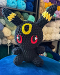 A black crocheted plush toy resembling a small fantasy creature with yellow ear patches, red circular eyes with white heart centers, and a yellow ring on the forehead, sitting on a blue surface with shelves of colorful yarn in the background.