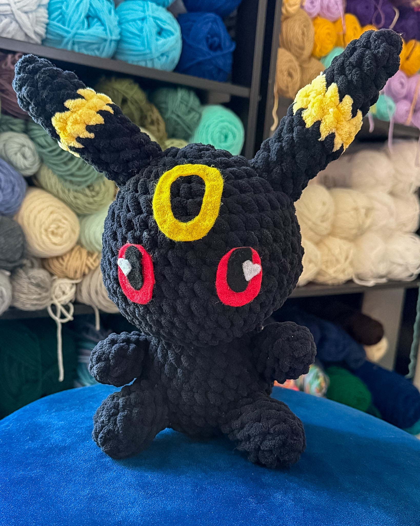 A black crocheted plush toy resembling a small fantasy creature with yellow ear patches, red circular eyes with white heart centers, and a yellow ring on the forehead, sitting on a blue surface with shelves of colorful yarn in the background.