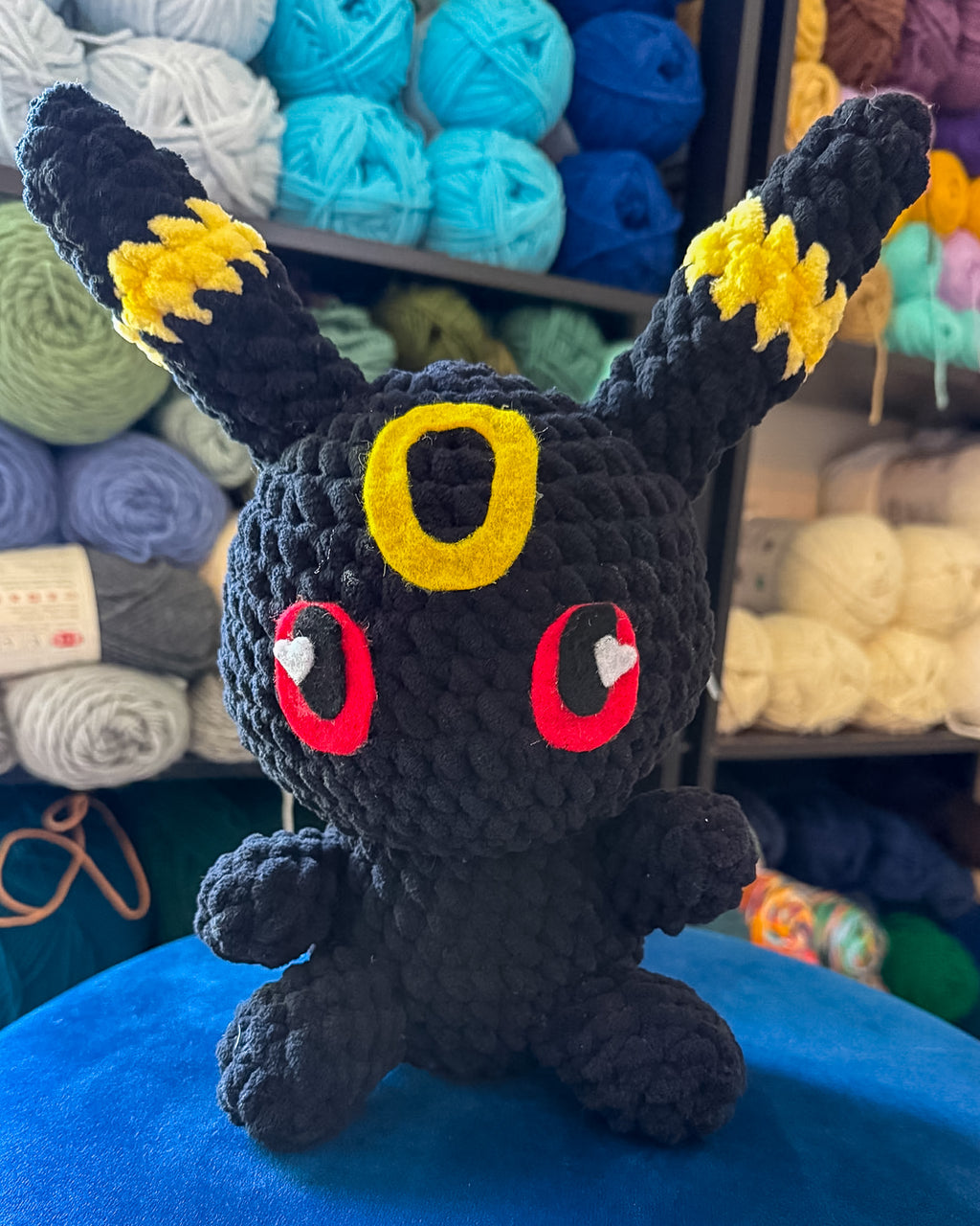 A handmade crocheted plush toy resembling a dark shadow creature with yellow ear tips, a yellow forehead emblem, and red eyes with white heart-shaped highlights, sitting on a blue surface with yarn balls in the background.
