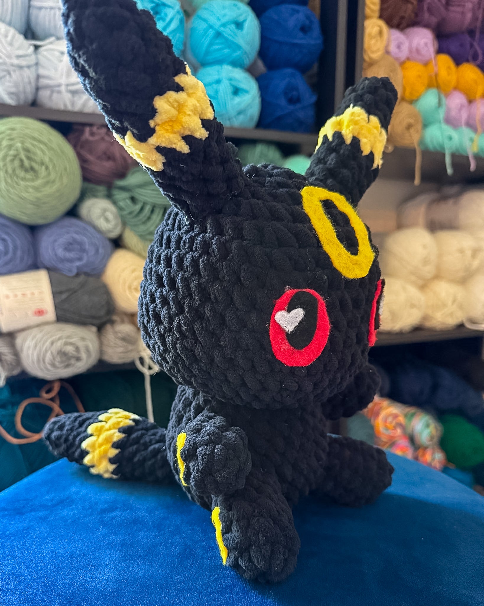 A handmade crocheted black Eevee plush toy with yellow, red, and white accents, sitting on a blue surface with shelves of colorful yarn balls in the background.