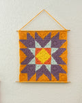 A square crocheted wall hanging with a geometric starburst pattern, suspended from a wooden dowel against a textured off-white wall. The design features purple, orange, yellow, and light blue sections.