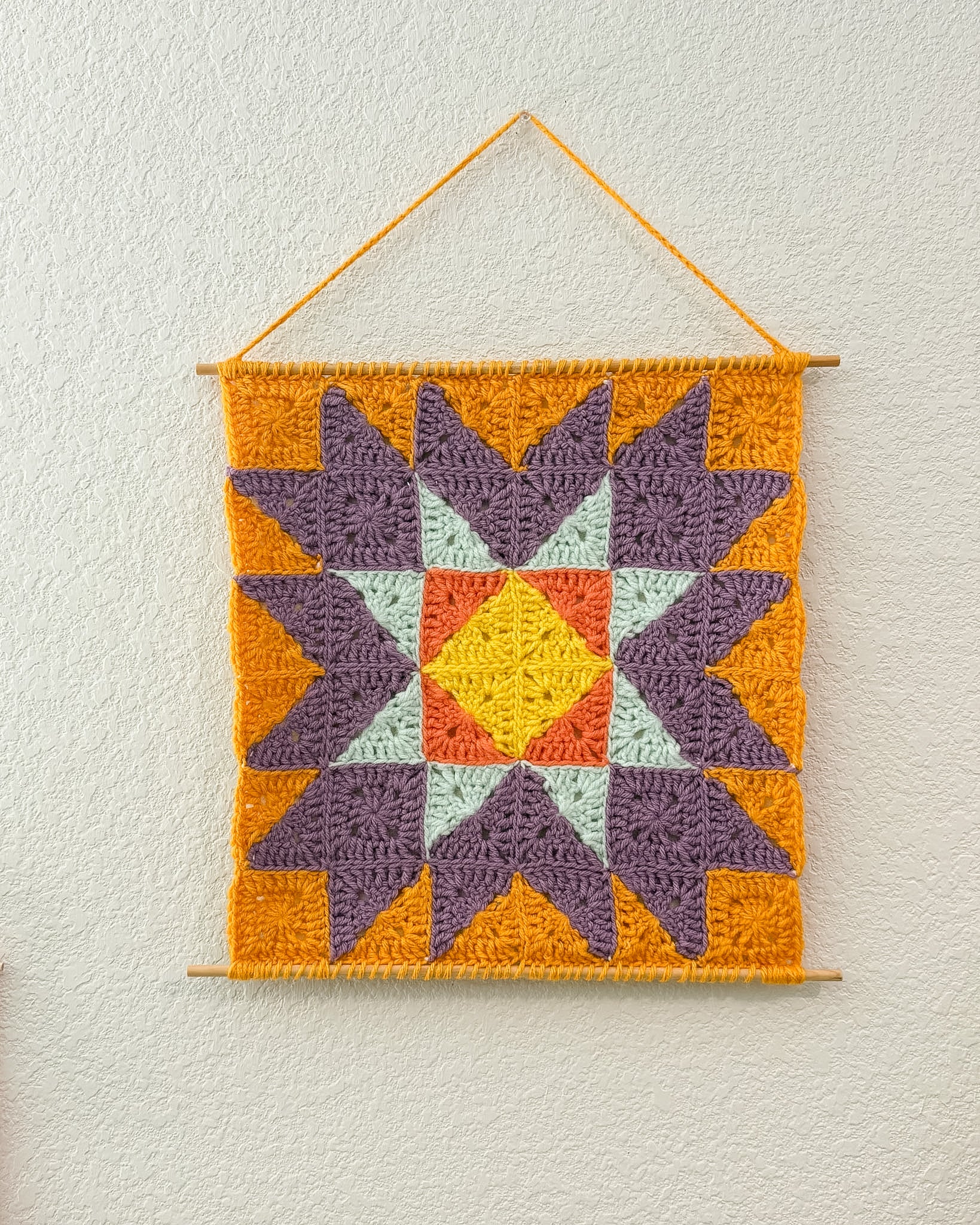 A square crocheted wall hanging with a geometric starburst pattern, suspended from a wooden dowel against a textured off-white wall. The design features purple, orange, yellow, and light blue sections.