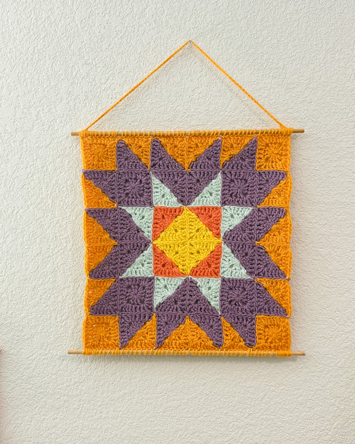 A square crocheted wall hanging with a geometric starburst pattern, suspended from a wooden dowel against a textured off-white wall. The design features purple, orange, yellow, and light blue sections.