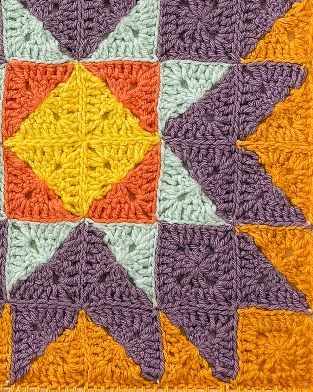 A square crocheted wall hanging with a geometric starburst pattern, suspended from a wooden dowel against a textured off-white wall. The design features purple, orange, yellow, and light blue sections.